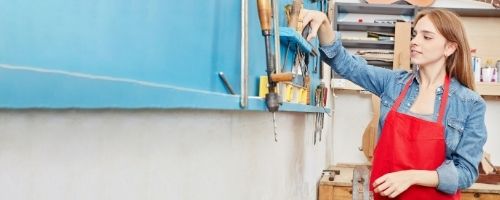 Female Apprentice In Workshop Wearing A Red Apron Looking At Tools 