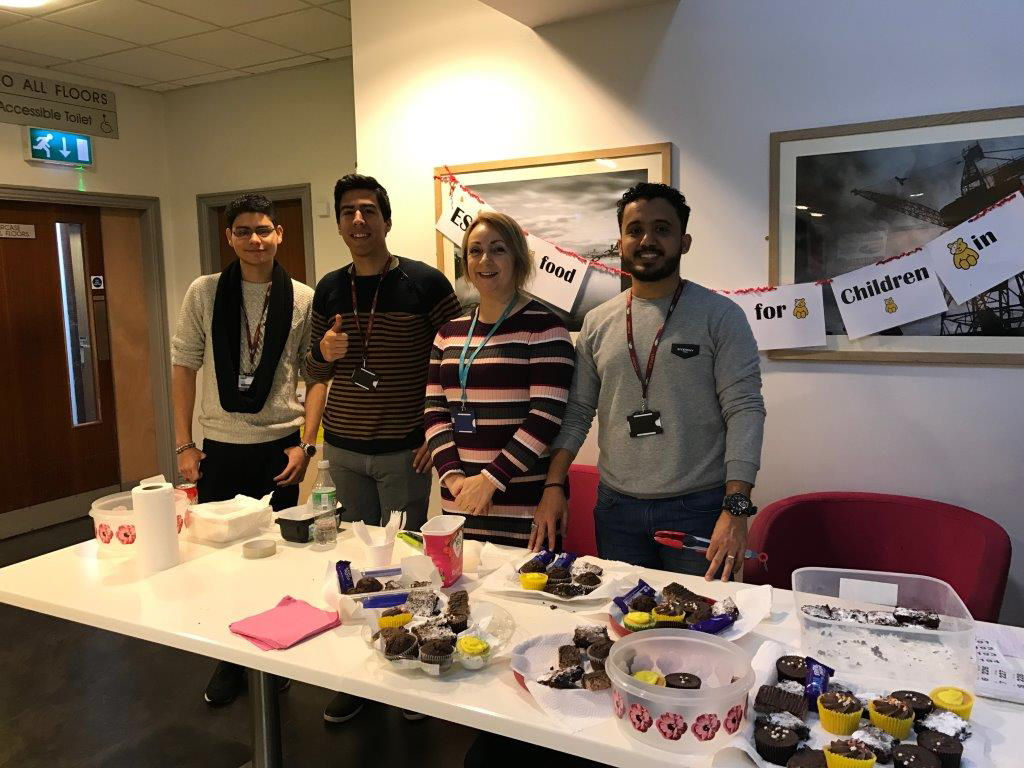 3 Wirral Met Students and 1 Staff Member standing by a table while volunteering for a Children in Need cake sale