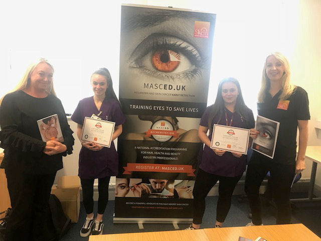 Wirral Met Beauty Students standing next to skin cancer charity pop-up banner with charity workers