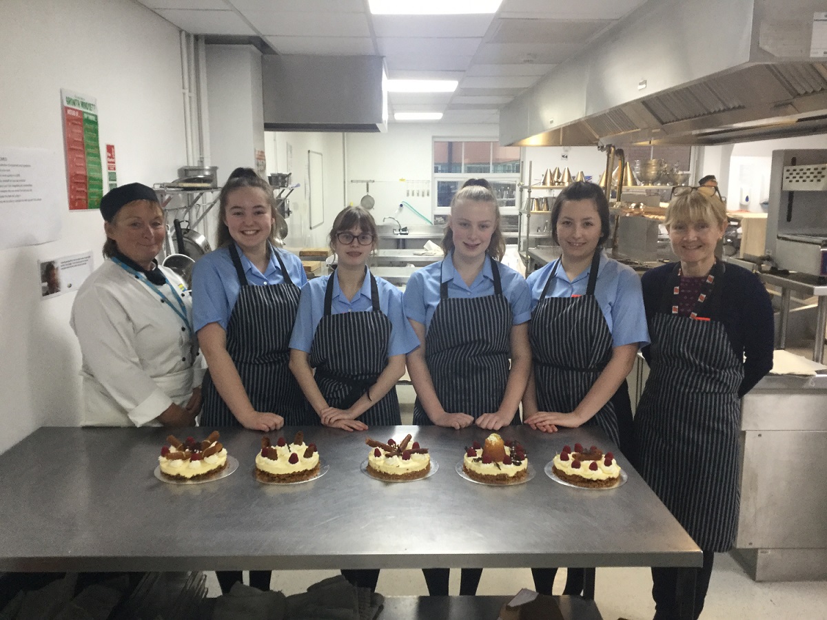 West Kirby Grammar School students standing in front of a table of cakes wearing aprons alongside teachers during Pastry Masterclass
