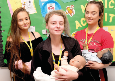 Three Wirral Met Childcare students standing and holding practise babies inside of a classroom