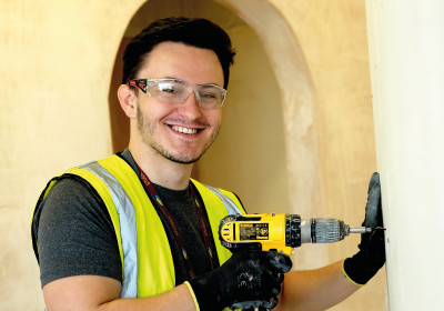 Male Part Time Construction Student Wearing Hi Vis Jacket And Drilling In To Wall