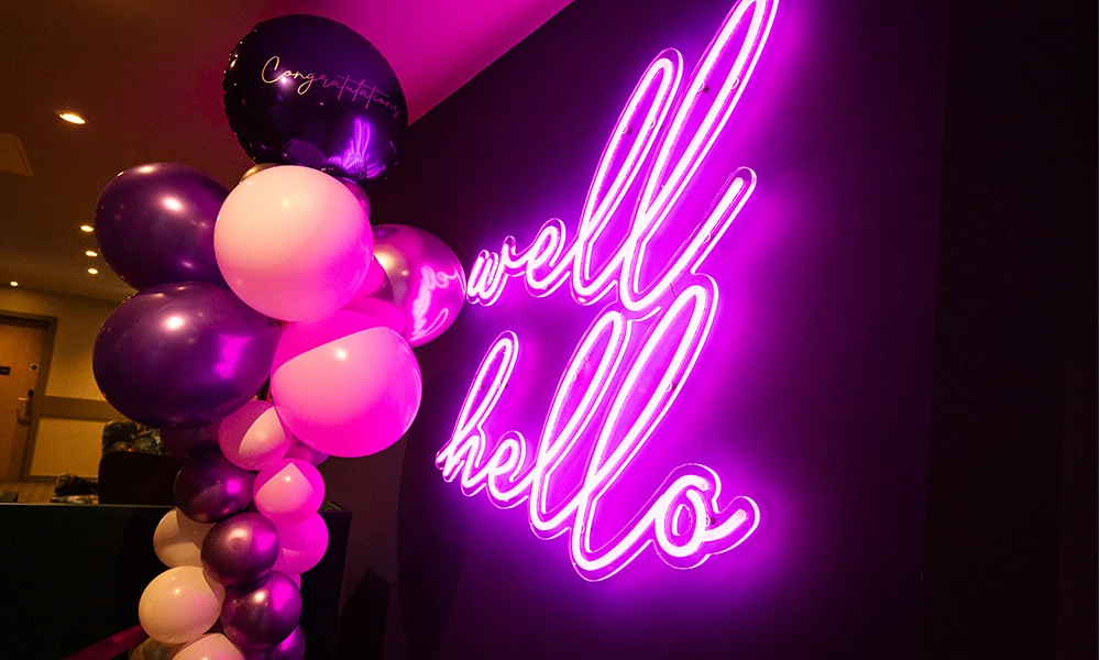 A neon purple sign saying well hello and baloons