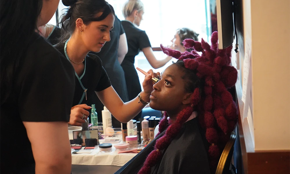 a female with red hair getting her makeup done by a female with a uniform on