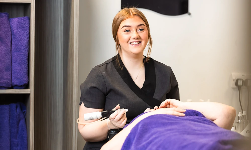 A female in a black tunic smiling at the camera with a lady on a bed for a beauty treatment