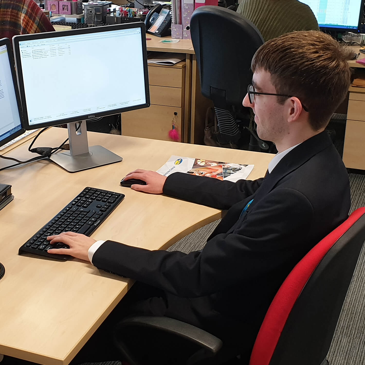 Man in suit sat on red chair at a desk looking at two computer screens